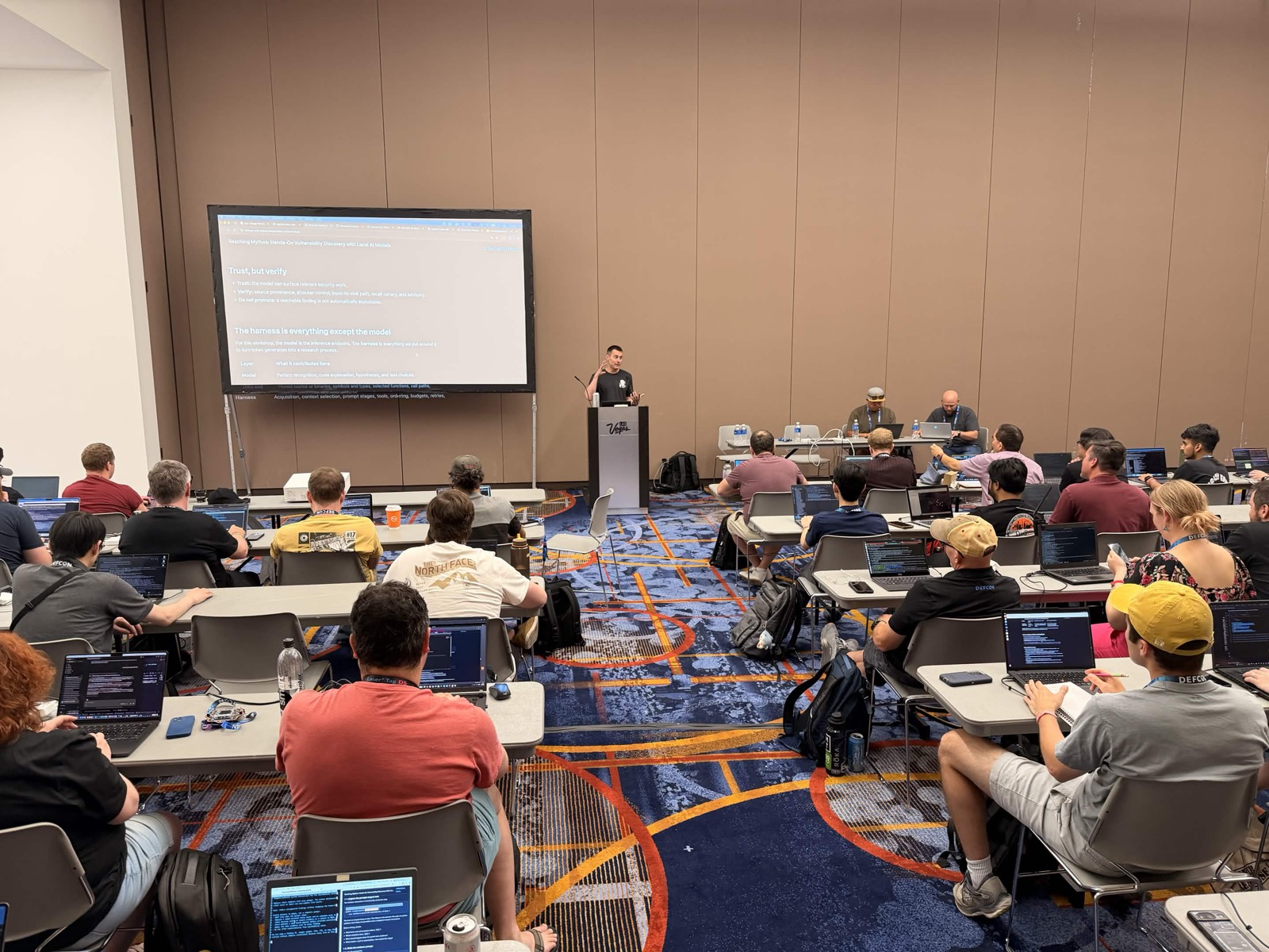 The Reaching Mythos workshop room at DEF CON 34, attendees on laptops with the Trust, but verify slide on the projector